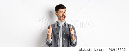 Funny handsome guy winking and pointing fingers at camera, inviting you check out promo, standing on white background 98918232