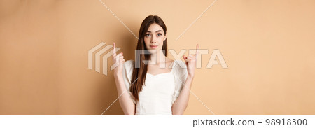 Hopeful romantic girl in white dress pointing fingers up, showing empty space for company logo, standing on beige background Hopeful romantic girl in white dress pointing fingers up, showing empty space for company logo, standing on beige background 98918300