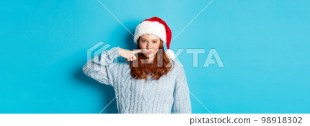 Winter holidays and Christmas Eve concept. Sassy teenage girl with red hair, wearing santa hat, enjoying New Year, poking her cheek and looking confident at camera Winter holidays and Christmas Eve concept. Sassy teenage girl with red hair, wearing santa hat, enjoying New Year, poking her cheek and looking confident at camera 98918302