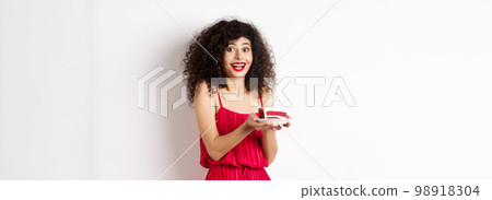 Birthday girl holding cake and making wish, smiling and celebrating, wearing festive red dress, white background 98918304