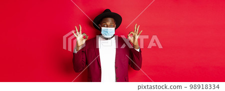 Covid-19 and fashion concept. Stylish african american man in hat and blazer, wearing face mask and showing okay sign, standing over red background 98918334