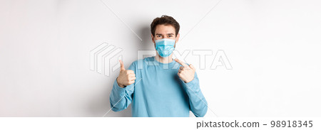 Coronavirus, health and quarantine concept. Smiling man pointing at his medical mask and showing thumb up, standing on white background 98918345
