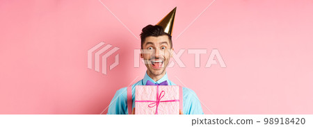 Holidays and celebration concept. Close-up of happy guy in party hat receive birthday gift, scream of joy and cheer, standing over pink background 98918420