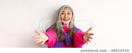 Close-up of stylish asian mature woman reaching hands forward, stretching arms for hug or hold something, smiling happy, standing over white background 98918439