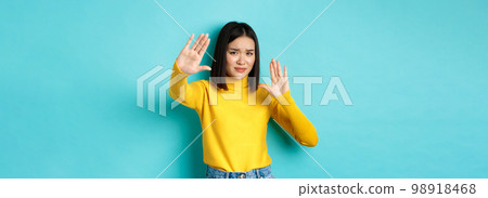 Image of woman feeling uncomfortable in front of camera, cover face with hands and frowning, begging to stop, standing against blue background Image of woman feeling uncomfortable in front of camera, cover face with hands and frowning, begging to stop, standing against blue background 98918468