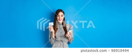 Online shopping. Beautiful woman paying for order with smartphone and plastic credit card, smiling at camera, wearing dress, blue background 98918494