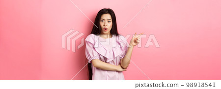 Shocked asian teen girl in dress pointing finger left, gasping wondered and look curious at camera, asking question about promo, standing on pink background 98918541