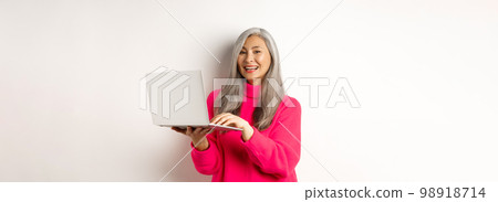 Beautiful asian senior woman entrepreneur working with laptop, laughing and smiling at camera, standing over white background 98918714