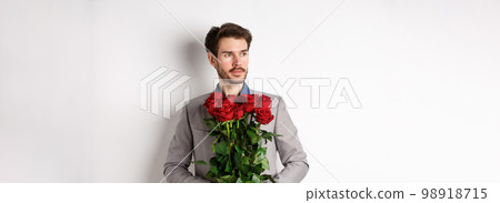 Handsome boyfriend in suit going on romantic date, holding bouquet of red roses and looking left thoughtful, standing over white background 98918715