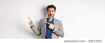 Excited handsome caucasian businessman in suit pointing right at logo, say wow and look amazed, standing on white background 98919361