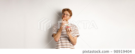 Holidays and celebration concept. Happy young man with red hair and glasses having birthday, holding cake with candle and thinking of b-day wish, standing over white background Holidays and celebration concept. Happy young man with red hair and glasses having birthday, holding cake with candle and thinking of b-day wish, standing over white background 98919404