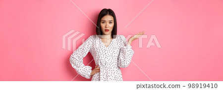 Careless asian woman raising hand and shrugging, staring at camera unbothered and questioned, so what gesture, standing over pink background 98919410