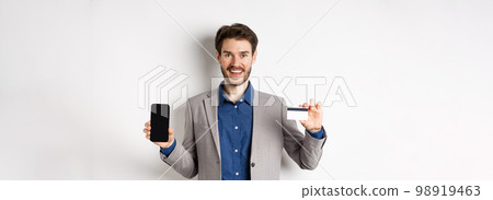 Online shopping. Smiling business man in suit showing plastic credit card with empty smartphone screen, standing against white background Online shopping. Smiling business man in suit showing plastic credit card with empty smartphone screen, standing against white background 98919463