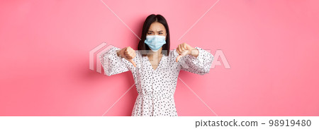 Coronavirus, social distancing and lifestyle concept. Disappointed asian woman in face mask looking judgemental, showing thumbs-down and disagree, standing over pink background Coronavirus, social distancing and lifestyle concept. Disappointed asian woman in face mask looking judgemental, showing thumbs-down and disagree, standing over pink background 98919480