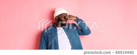 Disgusted african-american man shut nose from aversion, displeased by awful bad smell, standing in beanie and denim shirt over pink background Disgusted african-american man shut nose from aversion, displeased by awful bad smell, standing in beanie and denim shirt over pink background 98919490
