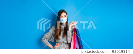 Covid-19, pandemic and lifestyle concept. Girl wearing medical mask on shopping in malls, holding paper bags and look aside at empty space, standing on blue background 98919514