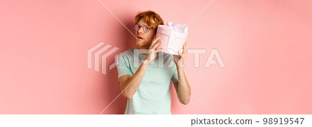 Valentines day and romance concept. Handsome redhead man shaking gift box and wonder what inside, trying to guess present, standing over pink background Valentines day and romance concept. Handsome redhead man shaking gift box and wonder what inside, trying to guess present, standing over pink background 98919547