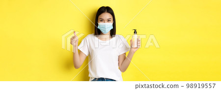 Covid, health care and pandemic concept. Asian girl in face mask from coronavirus, showing thumb up and good hand sanitizer, standing over yellow background Covid, health care and pandemic concept. Asian girl in face mask from coronavirus, showing thumb up and good hand sanitizer, standing over yellow background 98919557