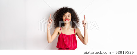 Smiling woman in elegant red dress and makeup, pointing fingers up and showing promo offer on valentines day, standing over white background Smiling woman in elegant red dress and makeup, pointing fingers up and showing promo offer on valentines day, standing over white background 98919559
