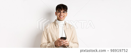 Real people. Handsome guy in glasses using mobile phone and smiling happy at camera, standing on white background 98919752
