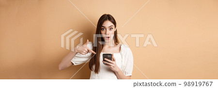 Online shopping concept. Excited girl pointing at smartphone screen and looking at camera amazed, standing against beige background 98919767