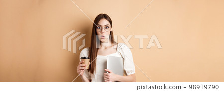 Working woman in glasses holding laptop and coffee, looking excited, hear amazing news, standing on beige background 98919790
