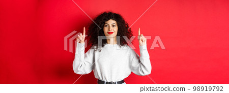 Image of cheerful smiling woman with curly hairstyle and red lips, pointing fingers up at empty space, showing advertisement, standing in casual clothes on studio background Image of cheerful smiling woman with curly hairstyle and red lips, pointing fingers up at empty space, showing advertisement, standing in casual clothes on studio background 98919792