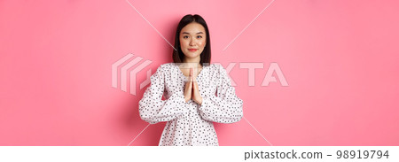 Beautiful asian lady in dress asking for help, holding hands in pray or namaste gesture, thanking you, standing over pink background 98919794