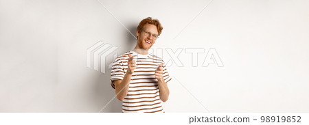 Cool and sassy redhead man with beard and glasses, pointing finger pistols at camera and smiling, congratulating you, standing over white background 98919852