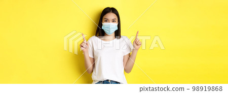 Covid-19, pandemic and social distancing concept. Young asian woman in white t-shirt and medical mask from coronavirus, pointing fingers up, showing special offer Covid-19, pandemic and social distancing concept. Young asian woman in white t-shirt and medical mask from coronavirus, pointing fingers up, showing special offer 98919868