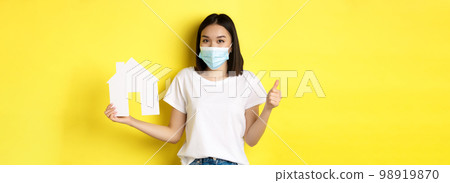 Covid-19, pandemic and real estate concept. Pretty asian woman smiling in medical mask, showing paper house cutout and thumb up, approve and like agency for buying property 98919870