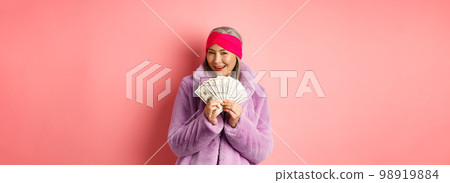 Shopping and fashion concept. Fashionable asian senior woman thinking about buying new clothes, showing money in dollars and smiling greedy, pink background Shopping and fashion concept. Fashionable asian senior woman thinking about buying new clothes, showing money in dollars and smiling greedy, pink background 98919884