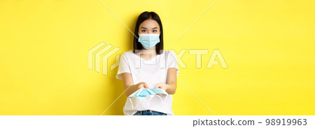 Coronavirus, quarantine and medicine concept. Cute asian girl giving medical masks and looking at camera, yellow background 98919963