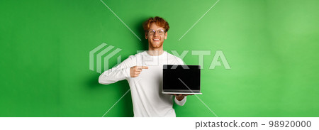 Happy redhead guy in glasses and white long-sleeve t-shirt, pointing finger at blank laptop screen and smiling, standing over green background 98920000