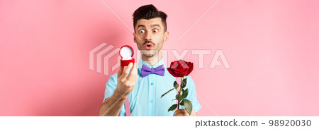 Valentines day. Romantic funny guy going to make wedding proposal, asking to marry him, holding red rose and looking at engagement ring with excitement, pink background Valentines day. Romantic funny guy going to make wedding proposal, asking to marry him, holding red rose and looking at engagement ring with excitement, pink background 98920030