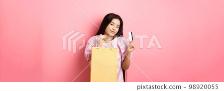 Shopping. Stylish asian girl showing shop bag and plastic credit card, standing on pink background 98920055