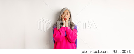 Surprised asian senior woman gasping amazed, holding hands on face and staring at camera impressed, standing over white background Surprised asian senior woman gasping amazed, holding hands on face and staring at camera impressed, standing over white background 98920058