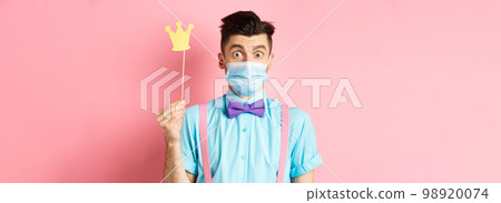 Covid, pandemic and quarantine concept. Cute and silly guy in medical mask looking surprised, holding small crown for party, standing over pink background Covid, pandemic and quarantine concept. Cute and silly guy in medical mask looking surprised, holding small crown for party, standing over pink background 98920074