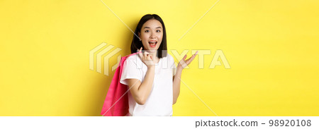 Cheerful asian female shopper looking amused, holding shopping bag and standing over yellow background Cheerful asian female shopper looking amused, holding shopping bag and standing over yellow background 98920108
