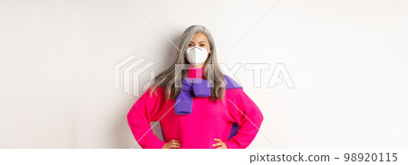 Covid, pandemic and social distancing concept. Confident and cheerful asian senior woman in respirator looking upbeat, smiling and holding hands on waist, white background 98920115