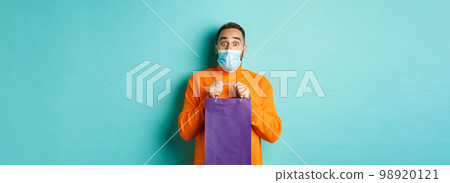 Covid-19, pandemic and lifestyle concept. Surprised man open shopping bag and looking amazed, receiving gift on holiday, standing over turquoise background 98920121