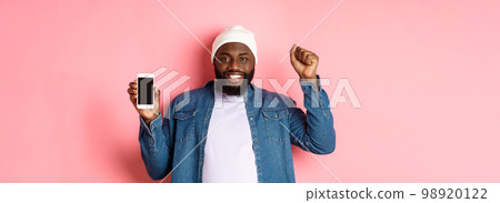Online shopping and technology concept. Cheerful Black man rejoicing and showing mobile screen, raising hand up satisfied, triumphing while standing over pink background 98920122