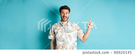 Surprised happy tourist pointing finger left at empty space, showing cool promo, standing in hawaiian shirt on blue background Surprised happy tourist pointing finger left at empty space, showing cool promo, standing in hawaiian shirt on blue background 98920145