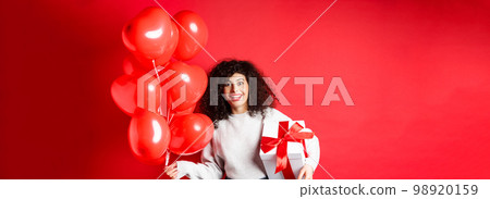 Excited beautiful woman celebrating Valentines day holiday, holding gift box and romantic heart balloons, looking surprised at camera, standing over red background 98920159