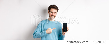 E-commerce and shopping concept. Smiling caucasian man pointing finger at empty smartphone screen, showing online offer, white background E-commerce and shopping concept. Smiling caucasian man pointing finger at empty smartphone screen, showing online offer, white background 98920168