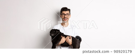 Happy hipster in glasses holding cute black pug and smiling, dog owner staring at camera with amazed smile, standing over white background 98920179