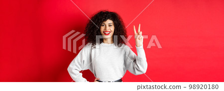 Young pretty lady in sweatshirt showing number two, making an order and smiling, standing on red background 98920180