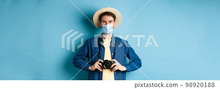 Covid-19, pandemic and travel concept. Young guy travelling abroad with camera, taking pictures on vacation, wearing medical mask from coronavirus, blue background Covid-19, pandemic and travel concept. Young guy travelling abroad with camera, taking pictures on vacation, wearing medical mask from coronavirus, blue background 98920188