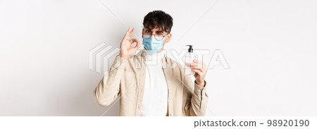 Health, covid and quarantine concept. Portrait of natural guy in glasses and face mask showing bottle of good hand sanitizer, make okay sign, recommending product, white background 98920190