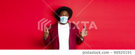 Covid-19, quarantine and holidays concept. Young african american man in medical mask, showing hand sanitizer and thumb up, recommending to use antiseptic 98920191